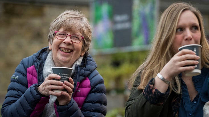 Two ladies in coats smile and laugh holding takeaway coffee cups